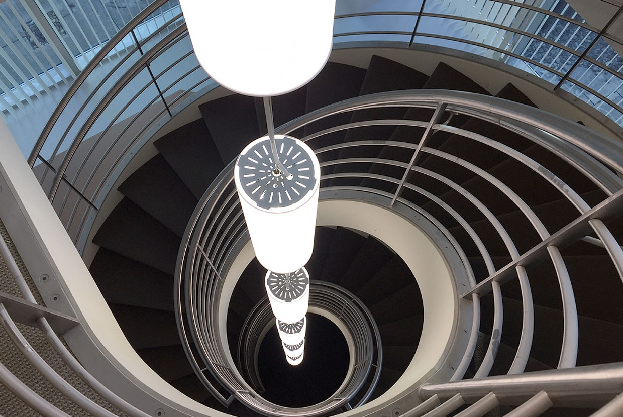 These clean, cylindrical custom light fixtures are hung in tandem down the center of round office stairwell