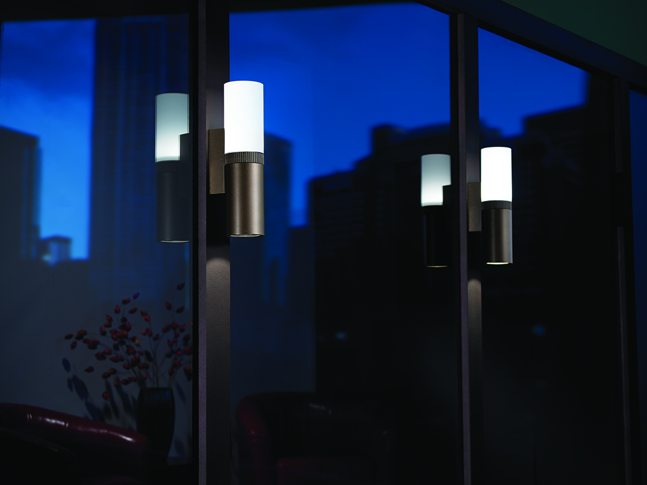 Scope exterior lighting fixtures illuminate large windows at night, reflecting a city skyline.