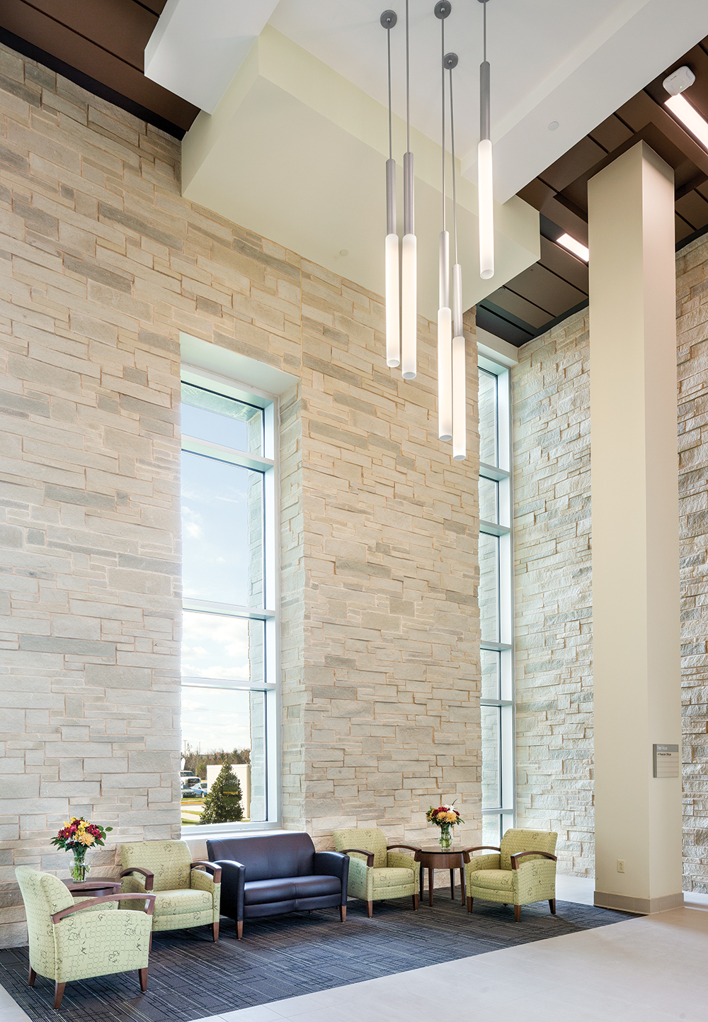 Sequence pendants highlight a clean, comforting healthcare design in a large waiting area with stone walls and green furniture.