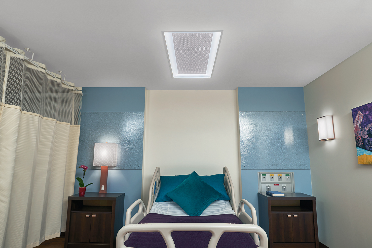 Serenity patient room lighting fixtures illuminate a hospital bed from a table lamp, overbed luminaire, and wall sconce.