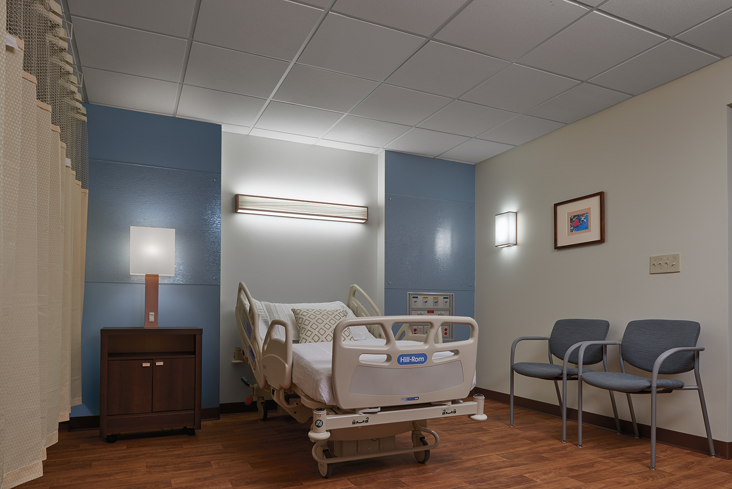 Serenity patient room lighting fixtures illuminate a hospital bed from a table lamp, headwall luminaire, and wall sconce.