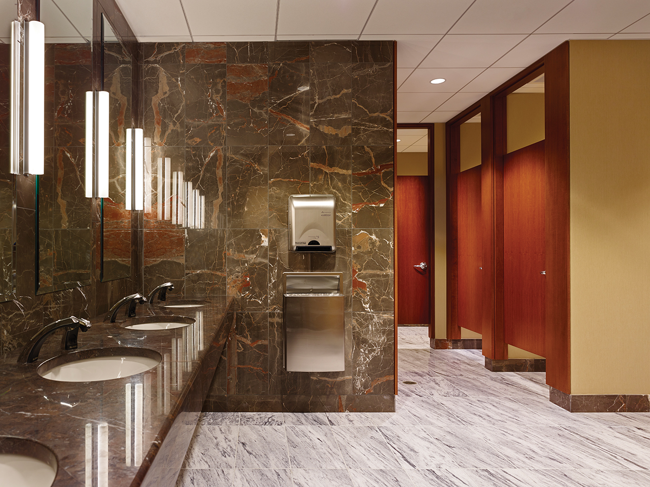 Voila modern vanity light fixtures illuminate mirrors in a public restroom with marble walls and countertops.