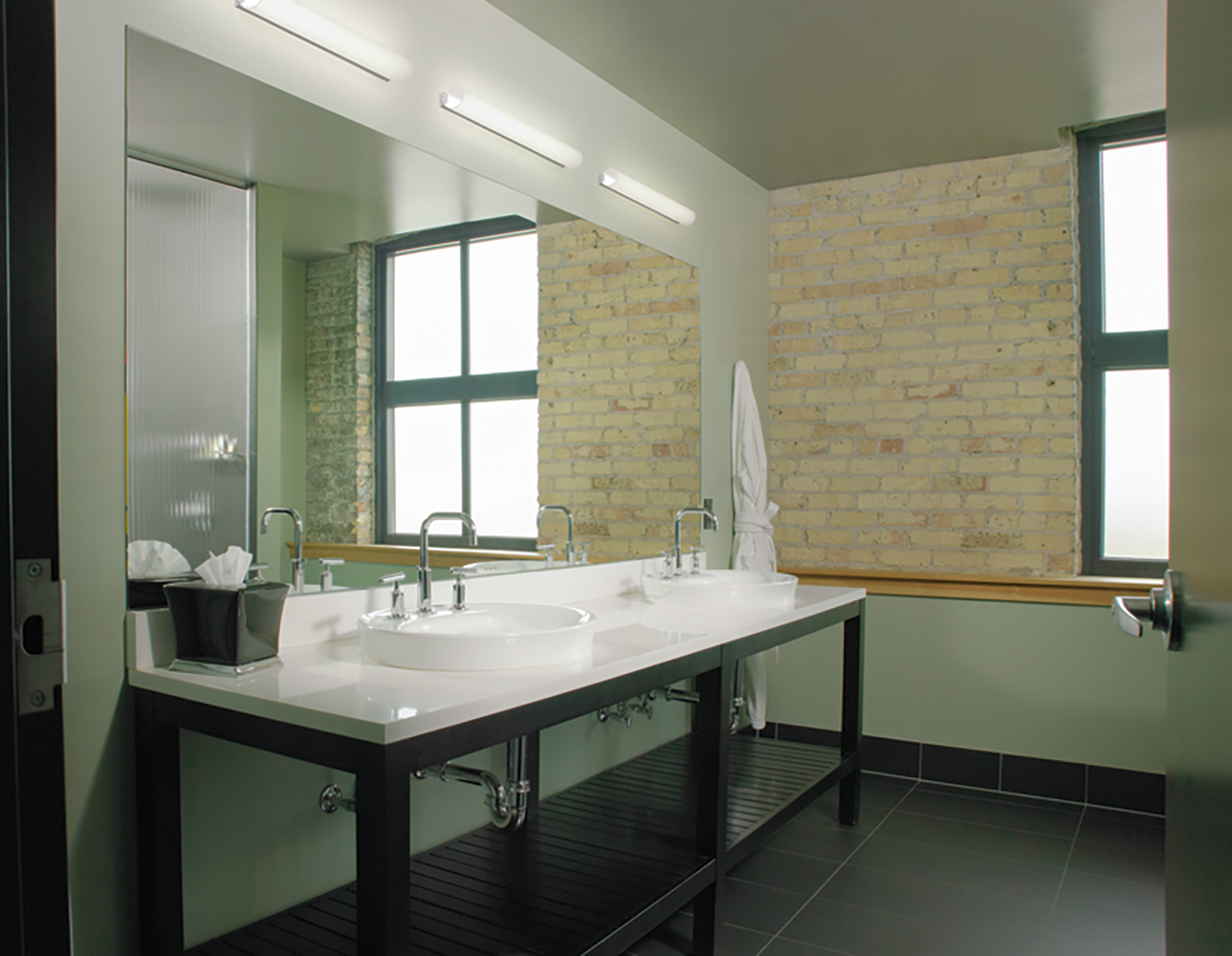 The Voila modern vanity light fixture, a luminous linear tube, above a clean mirror and double sink in a small bathroom.