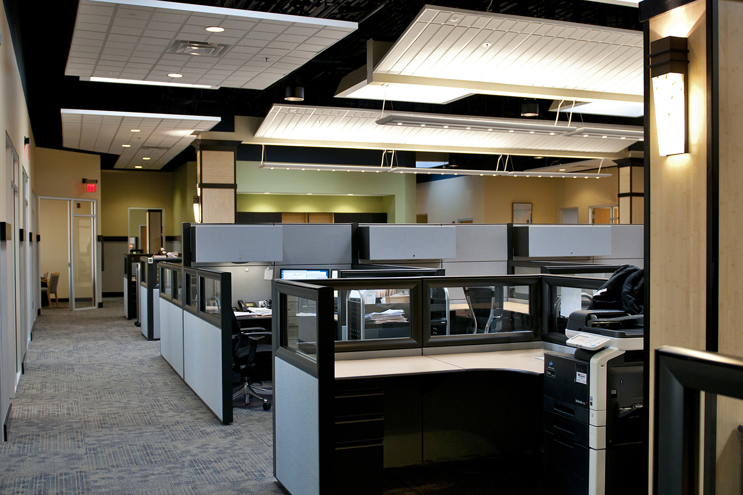 Wedge sconces illuminate a modern office lighting design, shown along the wall of an open cubicle area with an exposed ceiling.